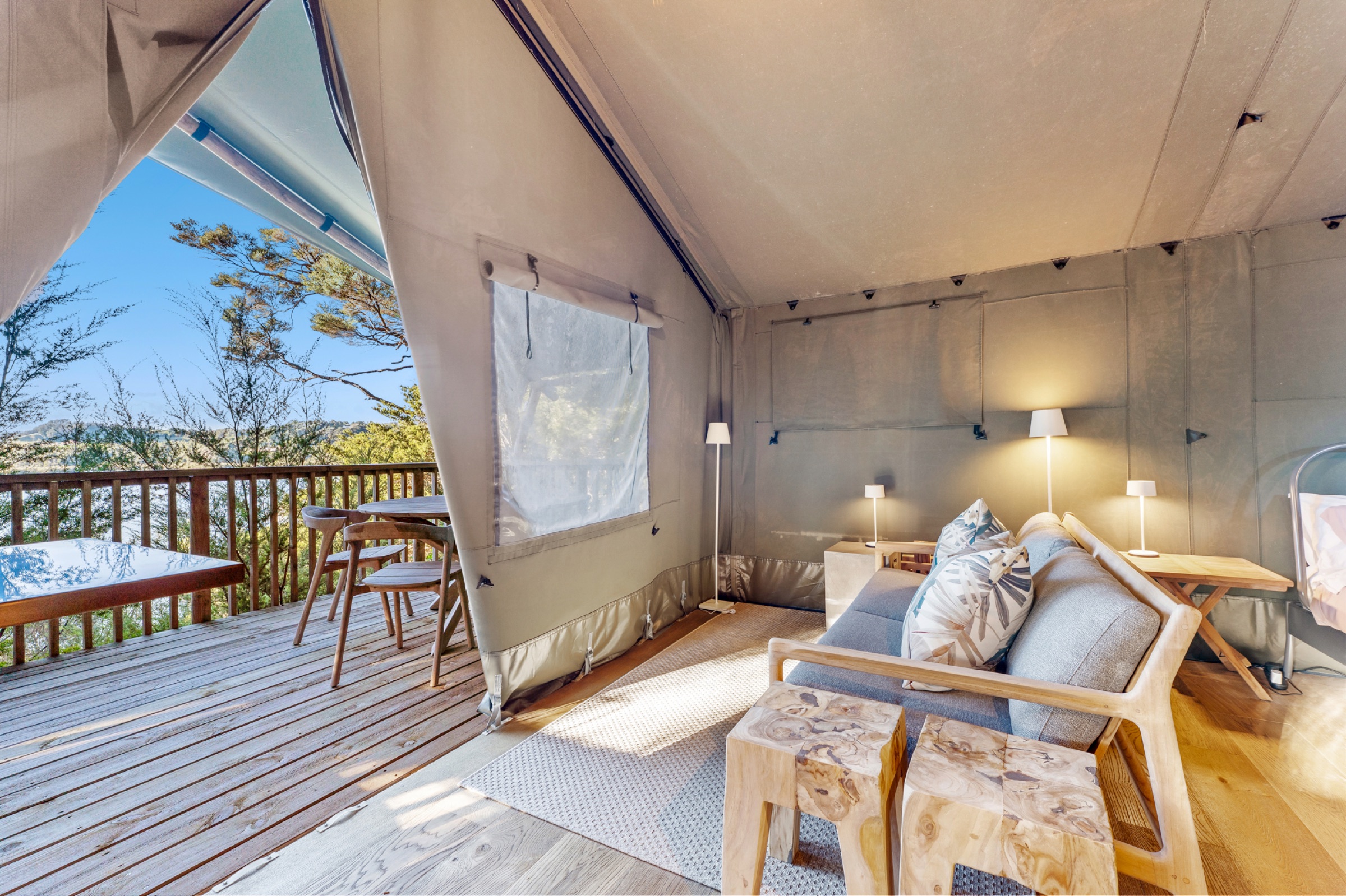 Inside Safari Tent 1 at Bluestone Encampment: a timber-framed sofa with grey cushions, two carved wooden stools, lamps on the side tables, and the canvas pulled back to the deck, the outdoor bath, and the inlet through the trees.