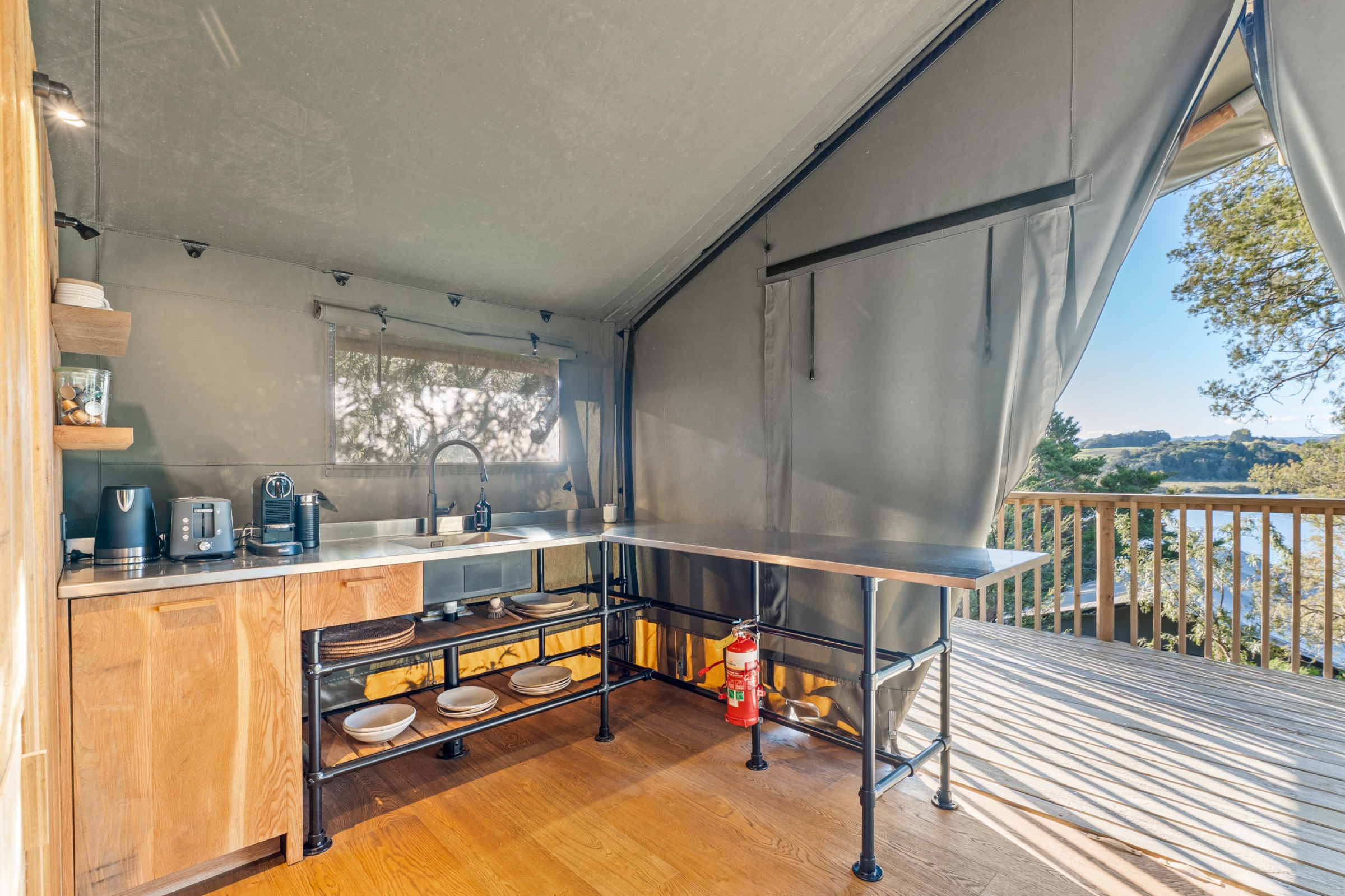 The kitchenette in Safari Tent 2 at Bluestone Encampment: a stainless bench with a sink, an espresso machine, a kettle and toaster, oak cabinetry, and the canvas rolled back to the deck and the inlet.