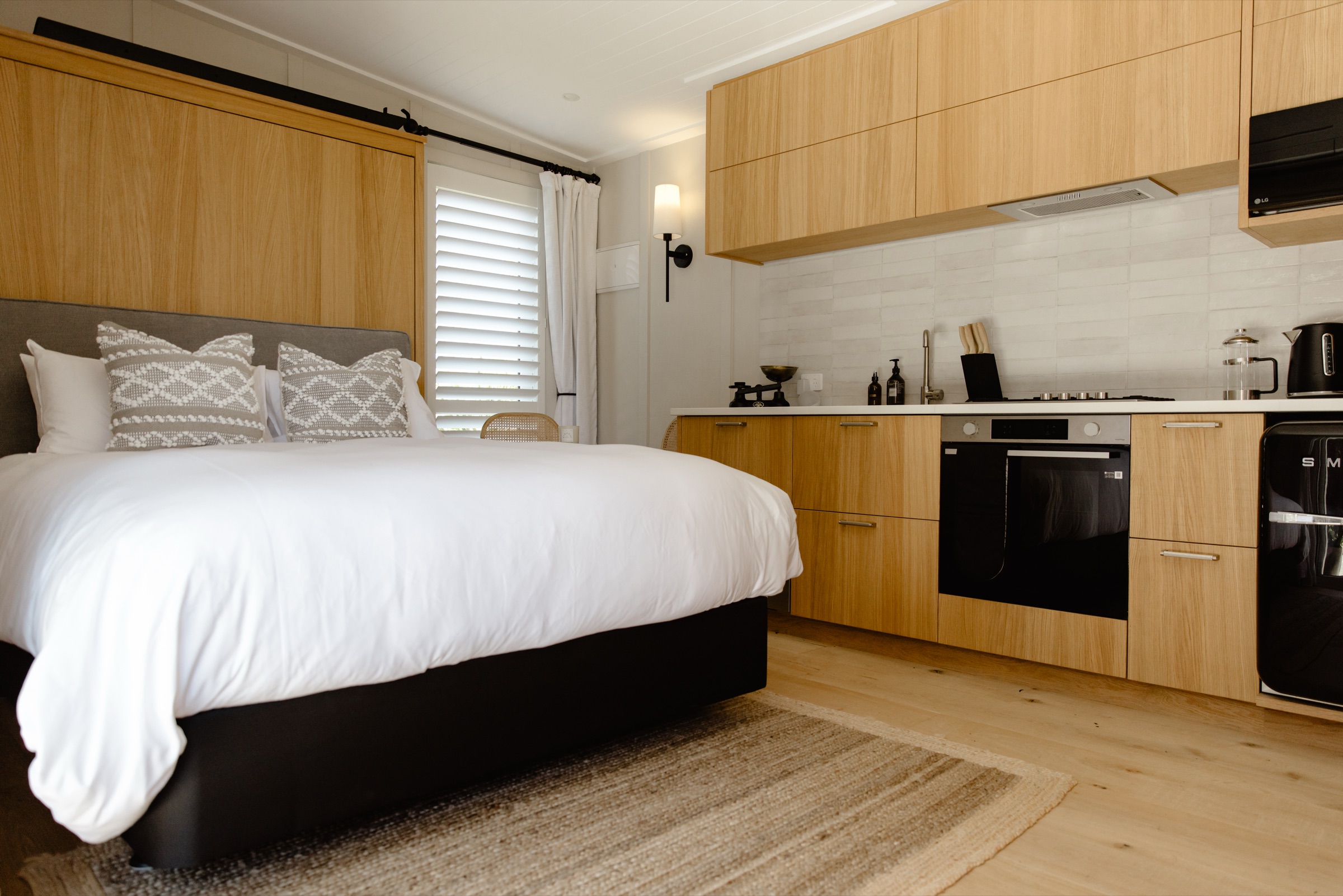 The Studio at Bluestone Estate in one frame: the queen bed with white linen and its oak surround, and the oak kitchen with a gas hob, an oven, and a fridge along the far wall.