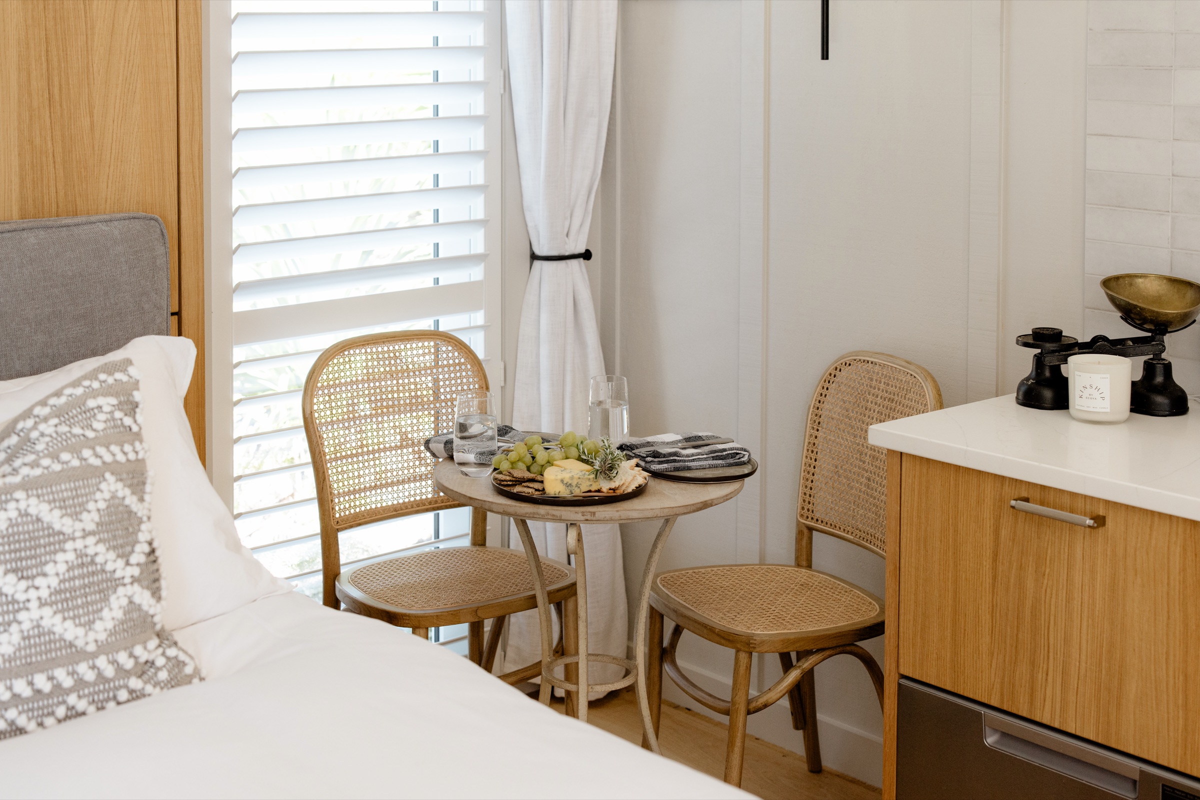 A cheese board and grapes on the small café table in the Studio at Bluestone Estate, two rattan chairs by the shuttered window, the bed beside and the oak kitchen bench with a candle and a set of brass scales.