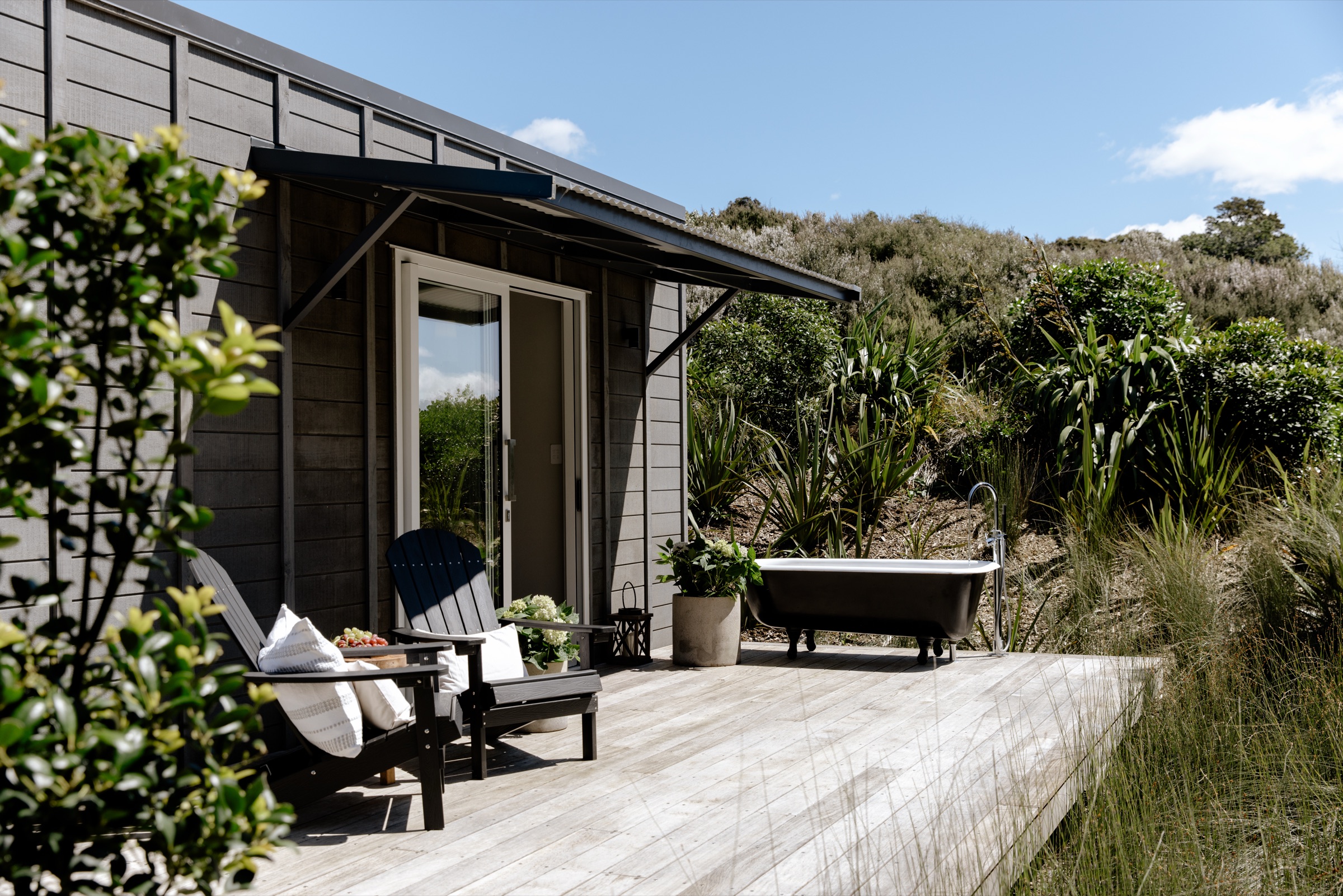 The Studio at Bluestone Estate: dark weatherboards, a sliding door onto the timber deck, two Adirondack chairs, and a freestanding bath outdoors among the flax and native planting.