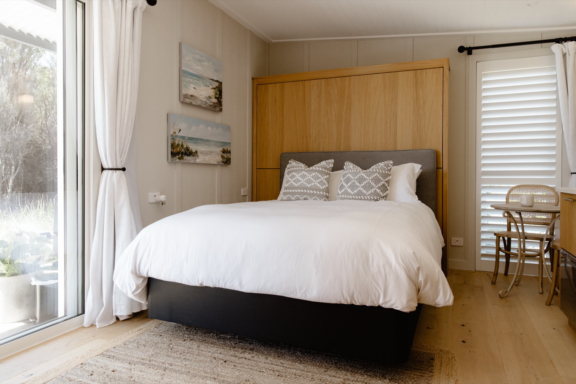 The Studio at Bluestone Estate: the queen bed with white linen against its oak wall panel, seascape paintings, linen curtains at the sliding door, and a small café table by the shuttered window.