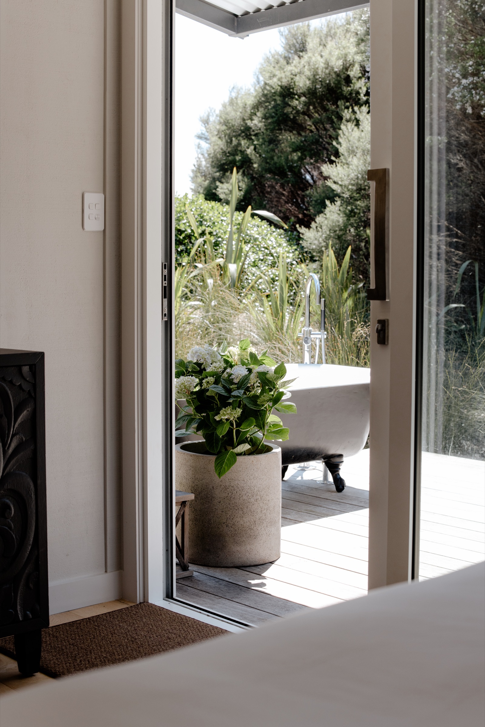 Looking out through the open sliding door of the Studio at Bluestone Estate: a white hydrangea in a concrete pot on the timber deck, the freestanding outdoor bath with its floor-standing tap, and flax and native bush beyond.