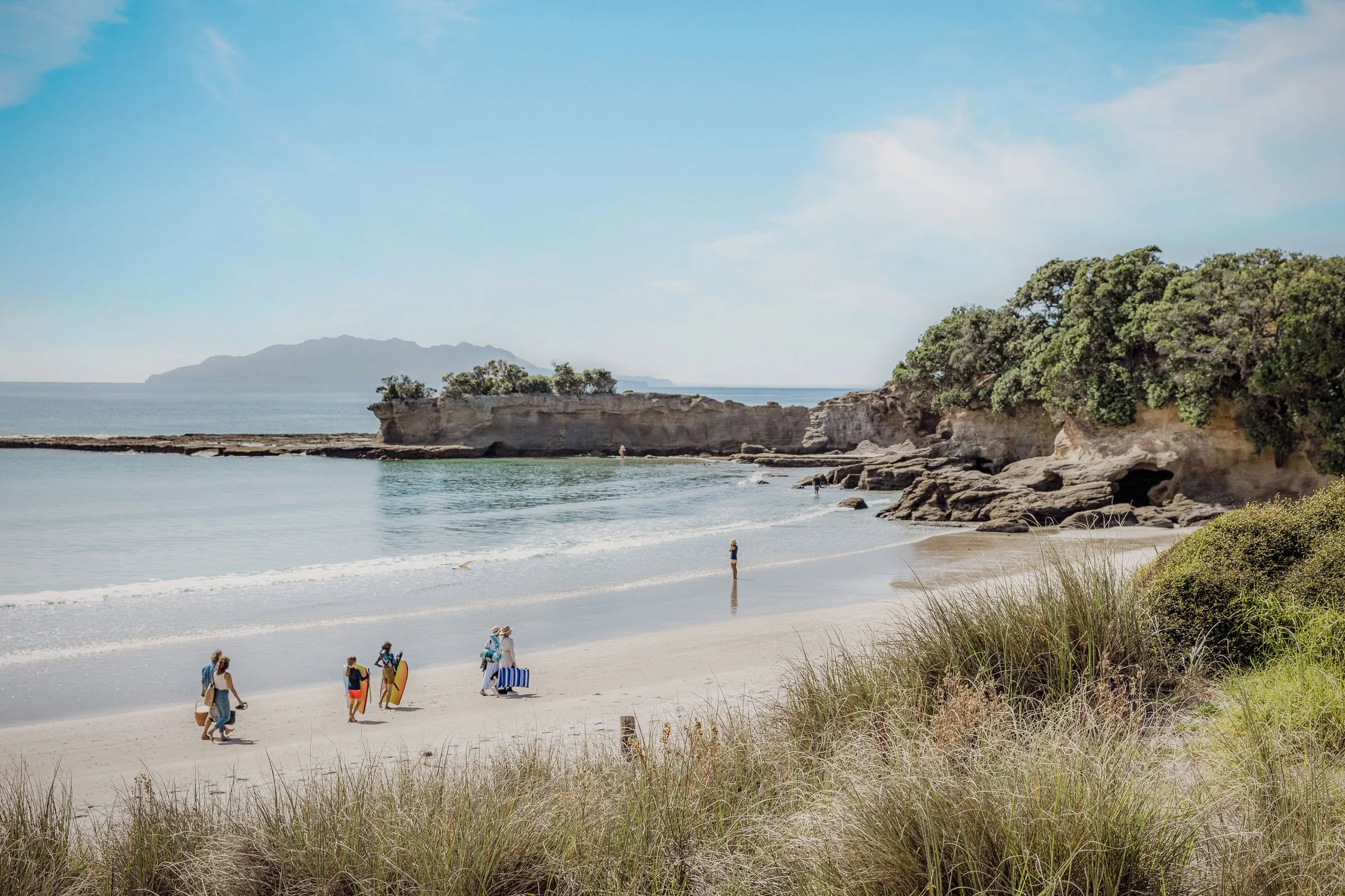 The white-sand beach at Tāwharanui Regional Park, with pōhutukawa on the headland and families walking to the water.