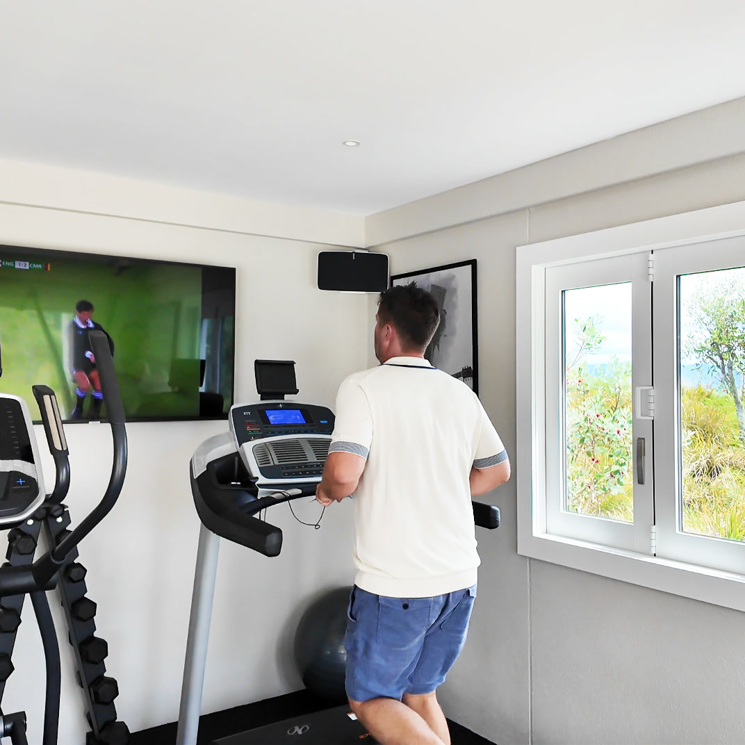 The Gym at Bluestone Main Estate: a guest on the treadmill beside the cross-trainer and dumbbells, the rugby on the wall screen, and the windows open to tussock, trees, and the hills.