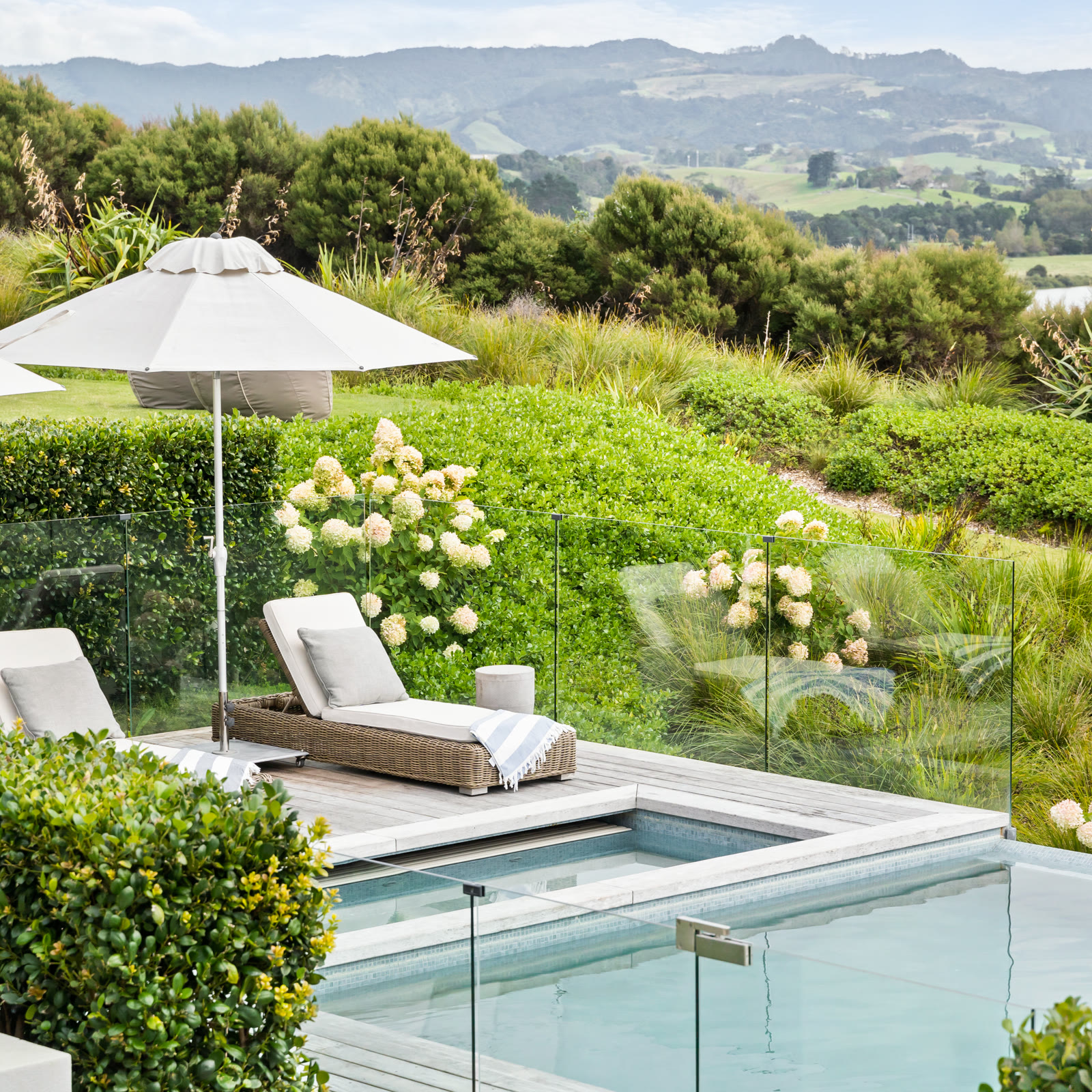 The pool and spa pool at Bluestone Main Estate: rattan loungers with striped towels under a cream umbrella on the deck, white hydrangeas along the glass fence, and the lawn, native planting, and estuary beyond.