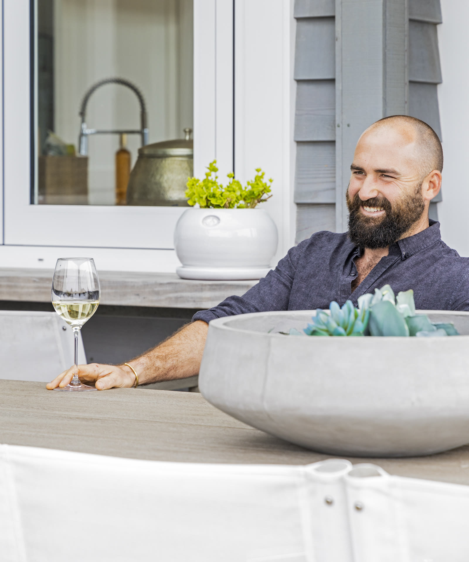 Will Simpson of Biome Gardens laughing at an outdoor table at Bluestone Estate, a glass of white wine in hand and a concrete bowl of succulents in front of him, grey shingles behind.